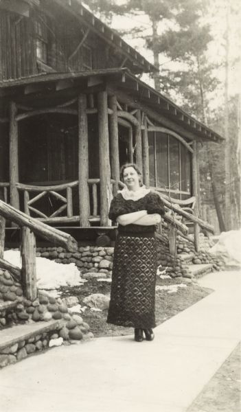 Woman standing on the sidewalk in front of the lodge at Trout Lake. The building is constructed of logs, with rustic railings, and stone slab steps with rock detail on the stairs.