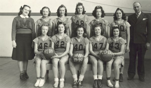 Group portrait of ten young women on a basketball team. Two women on the left are holding a basketball that has "Red's" written on it, and a woman in the center has a basketball with "Champs 39-40" on it. A woman is standing on the left wearing a letter sweater with an "M" on it. A man wearing a suit is standing on the right.