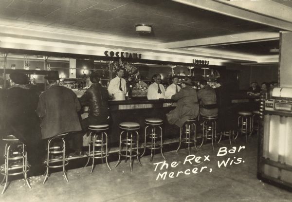People are sitting on stools along the bar, with bartenders behind serving drinks. Signs above the bar read: "Cocktails" and "Liquors." There is a jukebox along the right wall. Caption reads: "The Rex Bar, Mercer, Wis." Written on back: "Billy Goodreau, Howard Johnston."