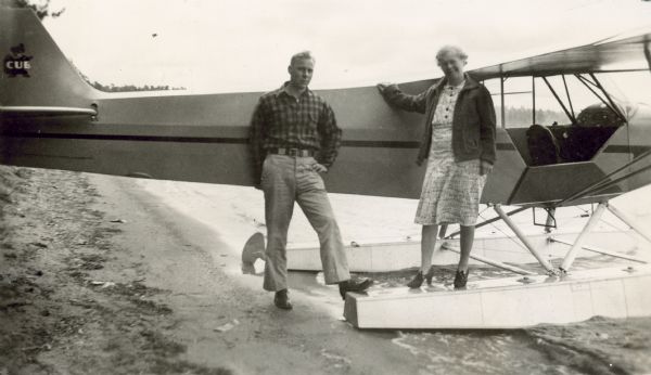 A man and a woman are standing on a shoreline in front of a fire watch seaplane pulled up onto the beach. On the tail of the plane is the Piper Cub logo featuring a bear, with the word "CUB" over it.