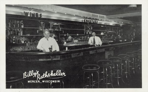 Two men are standing behind a bar. Above the back of the bar are signs that read: "Wines," "Cocktails" and "Liquors." Caption reads: "Billy's Rathskeller, Mercer, Wisconsin."