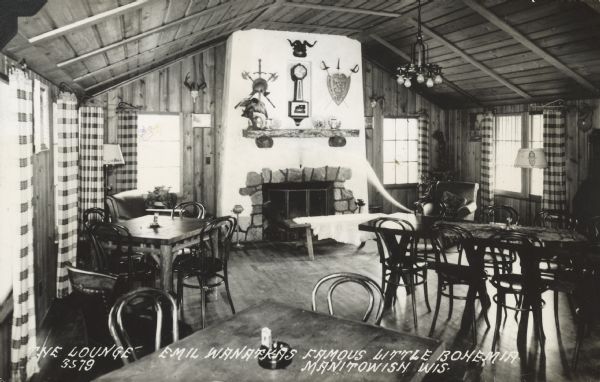 Interior view of the Lounge. A large fireplace is on the back wall. Armchairs, and chairs and tables are distributed around the room. Caption reads: "The Lounge, Emil Wanatka's Famous Little Bohemia, Manitowish, Wis."