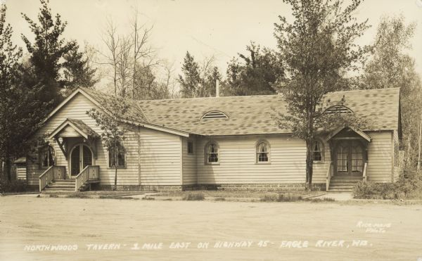 Exterior view of tavern, with two entrances. The windows and doors are arched at the top and there are eyebrow vents on the roof. Caption reads: "Northwoods Tavern — 1 mile east on Highway 45, Eagle River, Wis."