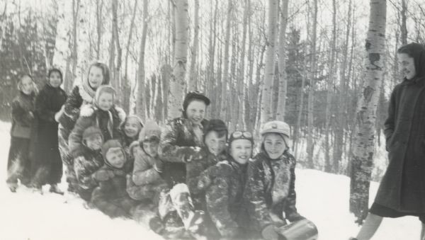 A large group of children are piled onto a toboggan. Other children are standing behind the toboggan on the left, and one young woman is standing on the right. Birch trees are in the background.