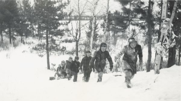View down hill toward a group of children walking up the hill in the snow. The children at the back of the line are dragging a toboggan. There appears to be a frozen lake at the bottom of the hill.