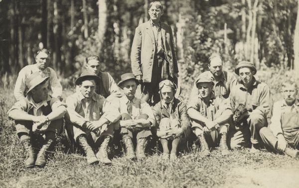 A group of men are posing outdoors. The man sitting in the center wearing goggles on his head is Jack Vilas, an air pilot (and forest fire spotter). A man on the right has a dog in his lap.