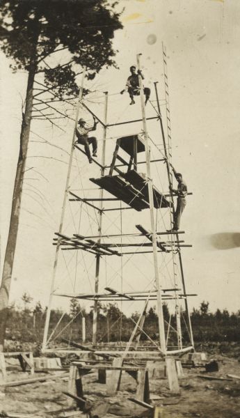Men are working up in the tower during construction. One man is standing mid-height on a ladder that runs the length of the tower on the right side. There is a fence and trees in the background.