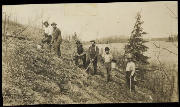A group of men are posing on the side of the hill. They are holding pails and other tools. A lake is in the background.
