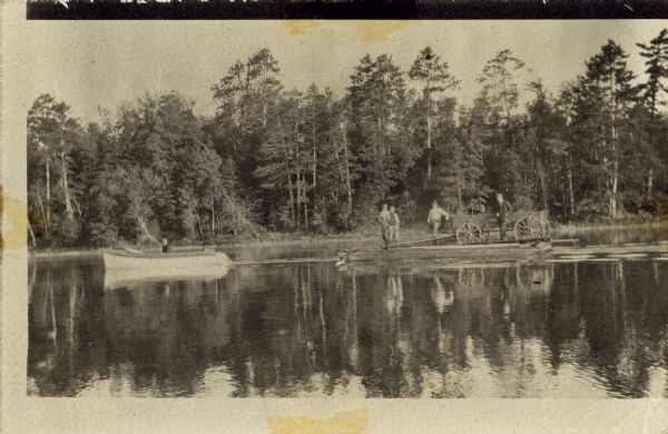 View across water towards a person in a boat on the left pulling a  group of men and a wagon on a raft. There is a tree-lined shoreline in the background.