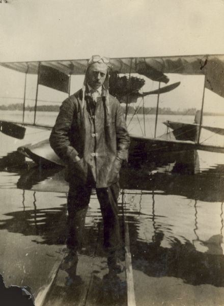 Jack Vilas standing on a pier partially submerged in the water. He is wearing a coat, pilot's cap, and goggles on his head. A seaplane is floating in the water behind him.