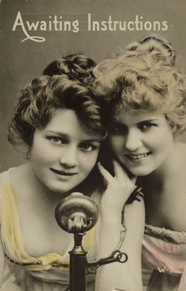 Hand-tinted photo postcard of two beautiful women, with their heads together, holding the receiver of a vintage telephone. They are wearing gowns and have their hair done elaborately. Text on front reads: "Awaiting Instructions."