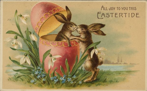 Easter postcard with two rabbits, one inside a hinged and decorated Easter egg, and the other rabbit outside. They are touching noses. The egg and rabbits are surrounded by white and blue flowers. In the background is a scene of a lake and trees. In the upper right corner is the text: "All Joy to You This Eastertide." Chromolithograph.