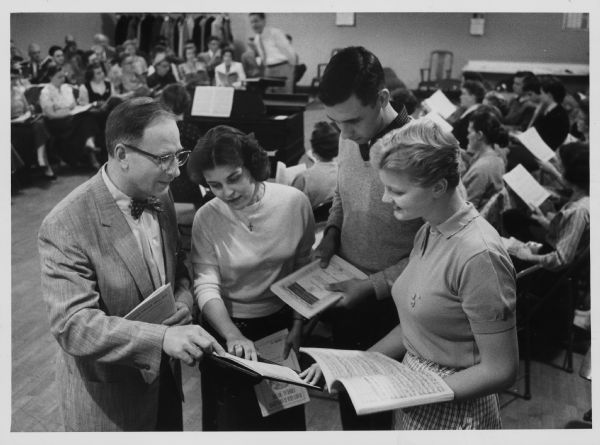 A man is pointing to sheet music on a stand, as a young man and two young women are looking on. Caption reads: "<b>The veteran Arion Musical club</b> chorus and the Carroll college chorus will combine at 3 p.m. Sunday to sing Brahms' 'A German Requiem' at the Milwaukee Auditorium. From left, Merrill Becker, Mrs. Robert Goralski, both Arions, Warren Johnson and Judith Johnson, Carroll students, look over their parts. The main groups are in the background. Dr. Lewis Whikehart of Carroll is director."