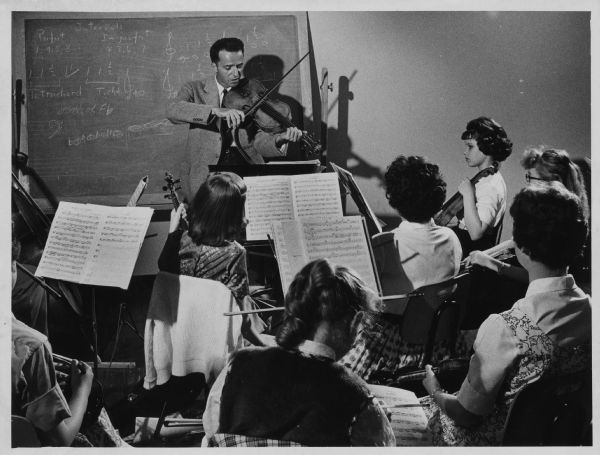 A man is playing a violin at the front of a classroom of students. Caption reads: "Rolf Persinger of the Chicago symphony orchestra worked Thursday with musicians in the Music for Youth program."