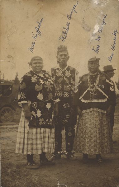 Outdoor group portrait of four Lac Courte Oreilles people: Mrs. [Lucille Mike] Kingfisher, Mitchell Quagon, with a pipe in his mouth, Lizzie Quagon, his wife, and Dan Homesky wearing a brimmed hat (in back). An automobile is parked in the background on the left.