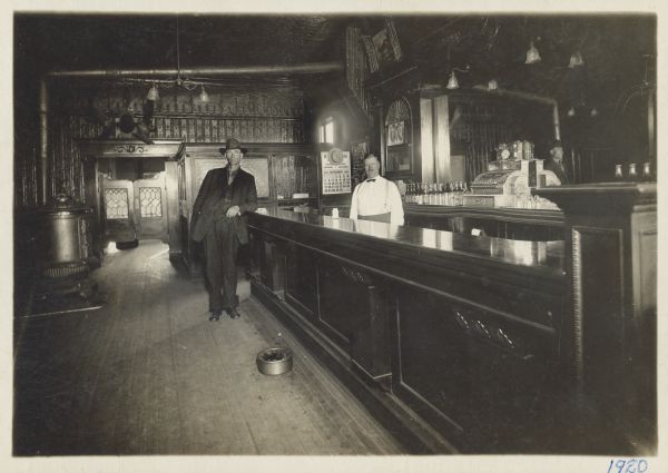 Two men are standing on either side of a saloon bar. The bartender on the right is identified as John Carlson. The bar has a stamped tin ceiling, and there is a large mirror behind the bar. In front of the mirror on the back bar counter is a large cash register with a clock on top. There is a spittoon on the floor in the foreground, and along the left wall is a woodburning stove. In the background the back wall is also of stamped tin, and there are saloon doors with a rack of antlers above it. A partitioned room with a pressed glass window and an opened door is in the right back corner, with a calendar on the right wall beside the door for September 1920.