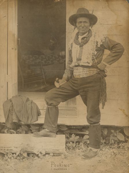 Outdoor portrait of a man standing outside the open door of a wooden home with one hand on his hip, and in his other hand holding what may be a pipe resting on his thigh. He is wearing a wide-brimmed hat and neckerchief, as well as a tasseled vest with epaulets and a belt. He is identified in a caption as "Posking" of the Chippewa (Ojibwe).