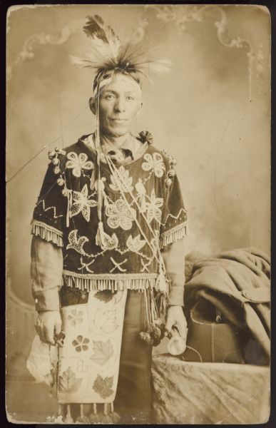 Studio portrait of a man who is a member of the Lac Courte Oreilles band of the Lake Superior Chippewa (Ojibwe). He is wearing a feathered headdress, a beaded vest/gibide'ebizon, and a bandolier bag/gashkibidaagan.