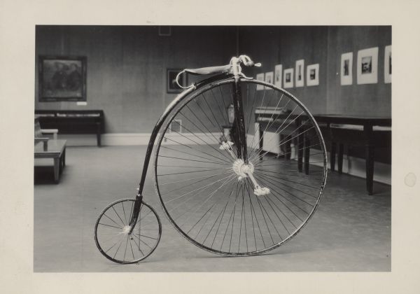 View of a penny-farthing bicycle, propped up indoors. Caption reads: "High wheel bicycle used by Dr. S. M. Babcock. H11560  Donor: S. M. Babcock estate."