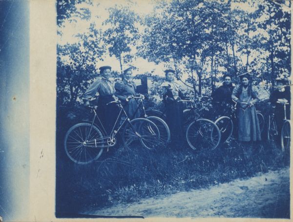 Group cyanotype portrait of six women posing with their bicycles in front of some trees. Caption reads: "Bicycle girls in a run near New London (?) about 1900."