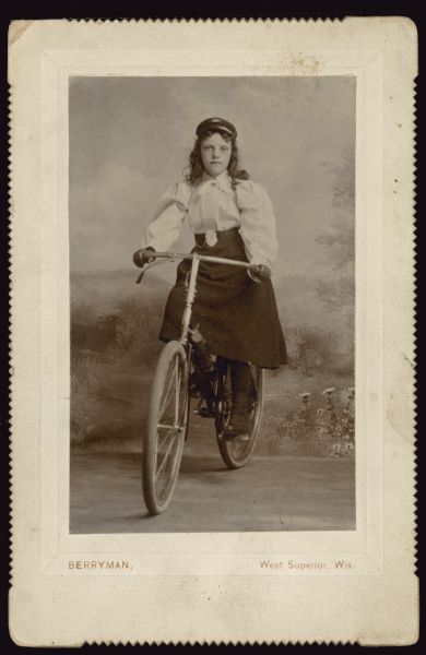 Carte-de-visite portrait in front of a painted backdrop of a girl on her bicycle. Caption reads: "11 years" and "Floy Montgomery."