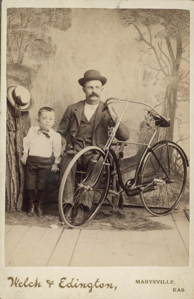 Carte-de-visite portrait in front of a painted backdrop of a man sitting in a chair and holding the handlebars of a bicycle, and a young boy standing next to him. There is a hat on top of a prop tree trunk on the left, and fake grass is on the floor. The man is holding on his knee what appears to be a stuffed monkey or other animal mounted on a board. Caption reads: "Comfort Wallace Young, My Dad. Gerald C. Young."