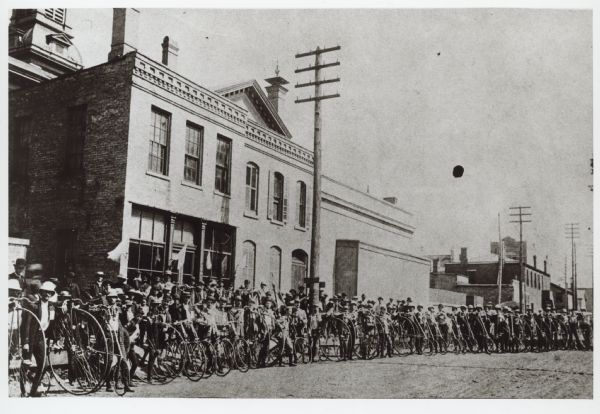A large group of people posing on a city street with bicycles. Caption reads: "Racine Bicycle Club ready for a run to Kenosha."