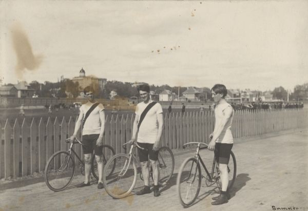 Three men posing with their bicycles along a picket fence. Caption reads: "On Camp Randall, 1899. Bicycle Team." The name "Sumner" is written in the lower right corner. There are horses and people in the field just beyond the fence. Bascom Hall is on the hill in the far background.