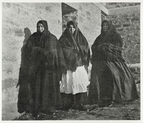 Group portrait of three women standing outside against a brick building. All three women are wearing dresses, with shawls covering their shoulders and heads. Caption reads: "Oneida women Copied from <u>The Church's Mission to the Oneidas</u> by Frank W. Merrill (Green Bay: News Publishing Co 1899) pg 4".
