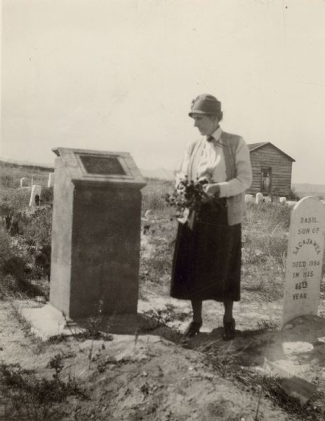 A woman holding flowers is standing between two graves, looking at the one on the left.
Caption reads: "Wind River Indian Reservation (formerly the Shoshone Indian Reservation), Fremont County, Wyoming. 1926. The grave of Sacajawea. The tablet on Sacajawea's monument reads: 'Sacajawea, Died April 9, 1884, a guide with the Lewis and Clark Expedition, 1805-1806. Identified 1908 by Rev. J. Roberts, who officiated at her burial.' Sacajawea died, in her sleep, of old age (about 100), at the Shoshone Agency, and was buried the same day, also on the Agency. 'In 1909, a concrete shaft with an imbedded bronze tablet was erected as a headstone for the grave of Sacajawea in the cemetery of the Shoshone reservation. The location of the grave was designated by the Reverend John Roberts, who officiated at the burial on [sic] Sacajawea on April 9, 1884. The shaft and bronze were donated by the Indian Agent, Mr. H. E. Wadsworth, and Mr. Timothy H. Burke.' From: Grace Raymond Hebard's, 'Sacajawea, Guide of the Lewis and Clark Expedition," The Arthur H. Clark Co., Glendale, 1933, page 298.
Basil was the adopted son of Sacajawea. He died in 1886, and was buried, Indian-fashion, in a creek bank. Remains disinterred to search for a wallet of L. & C. papers (found, but ruined), and reinterred January 12, 1925, beside Sacajawea.
'In 1932, two granite monuments were placed in the Shoshone Indian cemetery in memory of Basil, son of Sacajawea, and Barbara Baptiste Meyers, a daughter of Baptiste, and granddaughter of Sacajawea.' page 302.
Photograph presented by Grace Raymond Hebard of Laramie, Wyoming, who appears in the photograph."