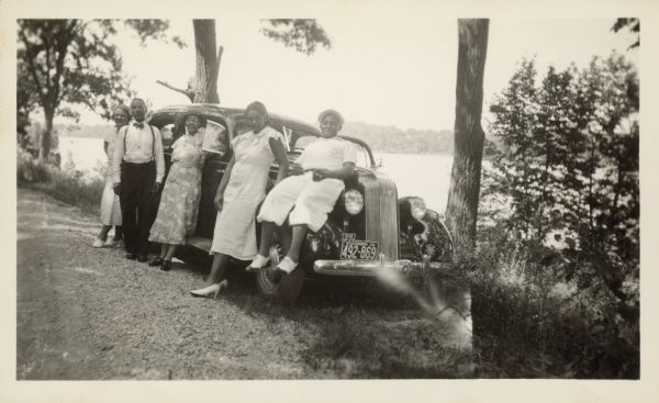 Three women and two men posing against a car. The car is parked near some trees and overlooking a body of water. A caption identifies four of the people as: "Louis Shepard, Julia Shepard, Bea Gulley, Carson Gulley," and identifies the location as "Pere Du Sac, Wisc."