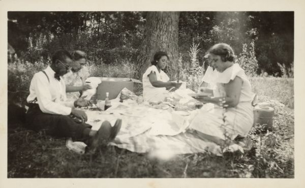 Three women and two men are sitting around a picnic lunch, which is placed on a cloth. A caption identifies three of the people as "Louis and Julia Shepard" and "Bea Gulley." Carson Gulley is also partly visible. 