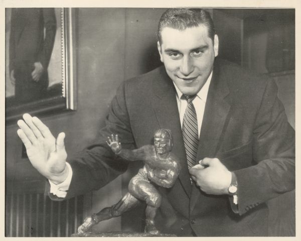 A man poses with a trophy. Caption reads: "NXP71-12/9-NEW YORK: Wisconsin fullback Alan Ameche assumes the same pose as the 20th annual Heisman Memorial Trophy which was awarded him by the Downtown Athletic Club of New York, 12/9. The trophy is awarded annually to the outstanding college football player of the U.S."