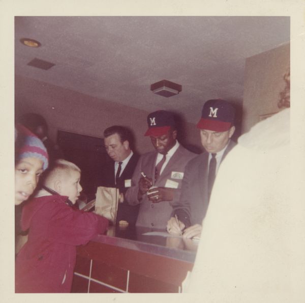 Three men are standing behind a counter, two of them wearing Milwaukee Braves baseball caps. In the middle, Hank Aaron is signing a baseball. To his left, another man is signing a document.