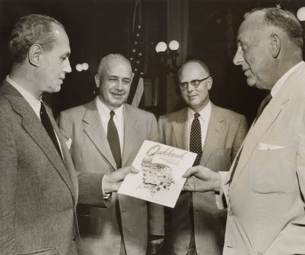Four men stand together, looking at a booklet entitled "Oshkosh: Wisconsin's City of Opportunity." They are (from left): Governor Walter Kohler, Connie Carver (Carver Ice Cream Co.), Leighton Hough (president of First National Bank of Oshkosh), and Assemblyman Harvey Abraham. 