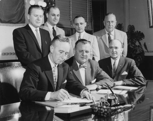 Three men are seated, and four are standing. Governor Vernon Thompson is signing Assembly Bill 26-A. Those present are: (Seated, left to right) Vernon W. Thompson; Lester S. Olsen; Clarence J. Muth; (standing left to right) Lester W. Brann; Assemblyman Earle E. Fricker; Assemblyman Glen E. Pommerening; Assemblyman Harvey R. Abraham.