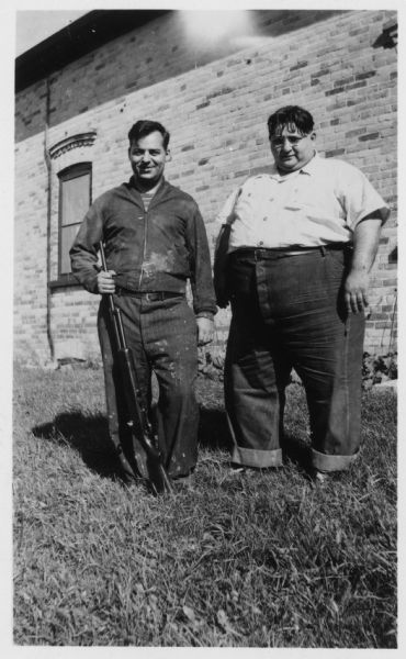 Two men pose outside a brick building. The man on the left is holding a rifle. Caption reads: "Thiensville (vicinity), Wis. Sept, 1943. Meyer Adelman and Emil Costello, on the farm they operated. Adelman was union organizer for SWOC [Steel Workers Organizing Committee] & 1st director of USWA [United Steel Workers of America] District 32. Costello was also SWOC organizer & 1st pres. (1937-1939) of Wis. State Industrial Union Council, & one-term Progressive Assemblyman (1937-1939)."