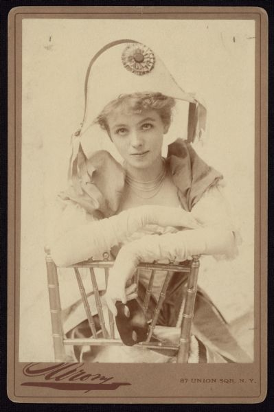 Seated carte-de-visite portrait of American stage actress Maude Adams. She is resting her arms on the chair back and is holding a mask in her hand. She is wearing a large hat, a dress with puffy sleeves and long gloves.