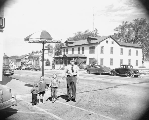 A uniformed police officer is holding hands with three children. They are standing in a crosswalk at the corner of Spring and Main streets. Behind them on the pole with the street signs is a large umbrella. Across the street is a large building with a sign that reads: "Hotel".