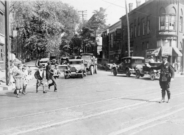 A uniformed police officer stands in an intersection and holds his hand out in the direction of several children. Cars are in the background. Caption reads: "Police Dept. Madison, Wis. Corner of Gilman & State Sts." According to Elroy Beckman, 1982: Officer Wilfred T. Schultz, about 1936. Corner State & Johnson.