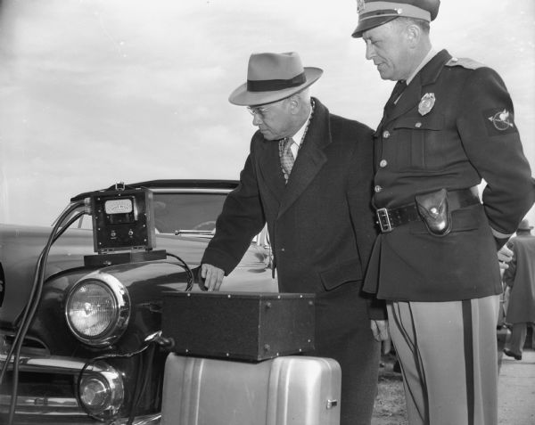 Two men are standing near a car, with the man on the right wearing a police uniform. A wired device is sitting on the hood of the car and both men are looking at it. Caption reads: "First police radar cars in Wis. Jefferson County." [A handwritten addendum follows] According to Elroy Beckman (ret'd Madison Police) Dane County ca. 1940. Judge Proctor & Ed Kelzenberg. 