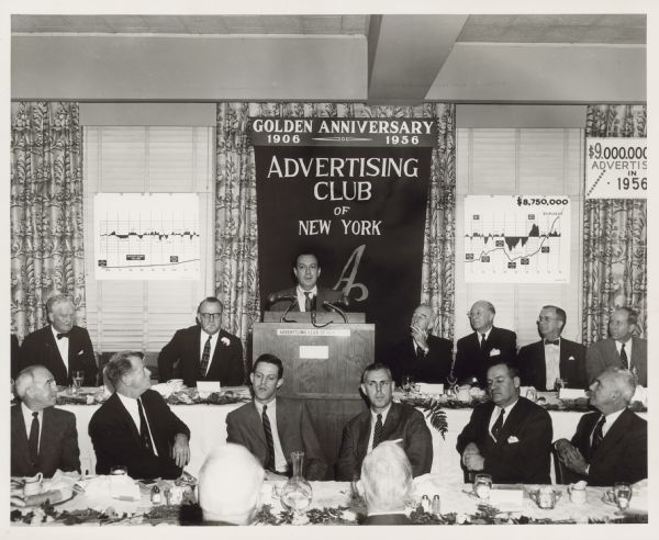 Several men are seated at tables while one man stands at a podium. Behind him a banner declares the Golden Anniversary (1906-1956) of the Advertising Club of New York.