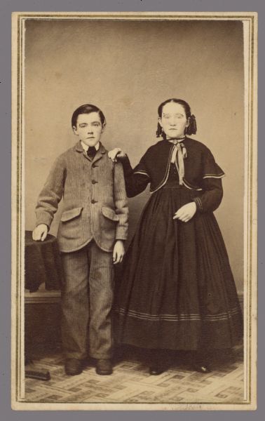 Full-length carte-de-visite portrait of a boy and a girl standing together. The girl has her hand on the boy's shoulder. These are the children of Joseph and Jane Ager, a farm family from Ripon.