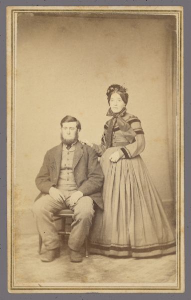 Carte-de-visite portrait of a man and a woman. Joseph Ager, a farmer from Ripon, is sitting. His wife is standing next to him with her hand on his shoulder.