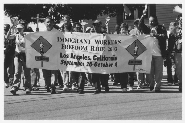 A group of people are marching on a street, holding small American flags, crosses, and a banner. The banner reads: "Immigrant Workers Freedom Ride 2003 Los Angeles, California September 20-October 4." Caption reads: "Immigrant Workers Freedom Ride comes through Morristown, Tennessee-October 2003."