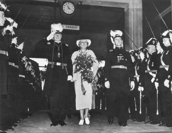 A woman and two uniformed men are walking together. They are passing between two rows of uniformed men presenting swords. Caption reads: "GRAND MASTER ARRIVES FOR CONCLAVE. MIAMI, Fla. - Andrew Davison Agnew (left) of Milwaukee, Wisc., accompanied by his wife, being saluted upon arriving here by special train to preside over the 40th Triennial Conclave of the Knights Templar, July 17 to 23. At right is John B. Phelps of Miami, general chairman of the national conclave."
