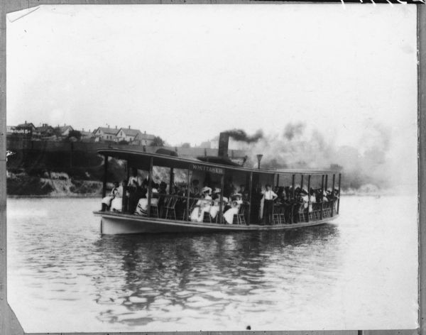 View of a boat, the <i>Whittaker</i>, with most people sitting in chairs, and a few men standing. In the background is a steep shoreline, with houses along the top behind a fence.