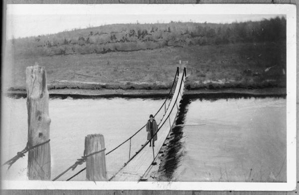 View looking down towards a young woman walking across a bridge. Caption reads: "Cable Bridge across Peshtigo River (Marinette County). Josephine Schaffer-one of those who cross every day in order to get an Education at the Kirby Hill School."