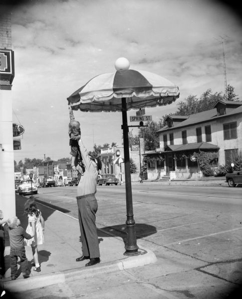 Caption reads: "Cambridge, Wis. Policeman holding child up to let her touch the umbrella on the "lamp post" which is the community's "trade-mark"."