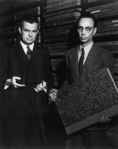 Portrait of Edward Alexander, director of the Wisconsin Historical Society, holding a reel of microfilm, and Benton Wilcox, librarian of the Wisconsin Historical Society, holding a bound newspaper volume.